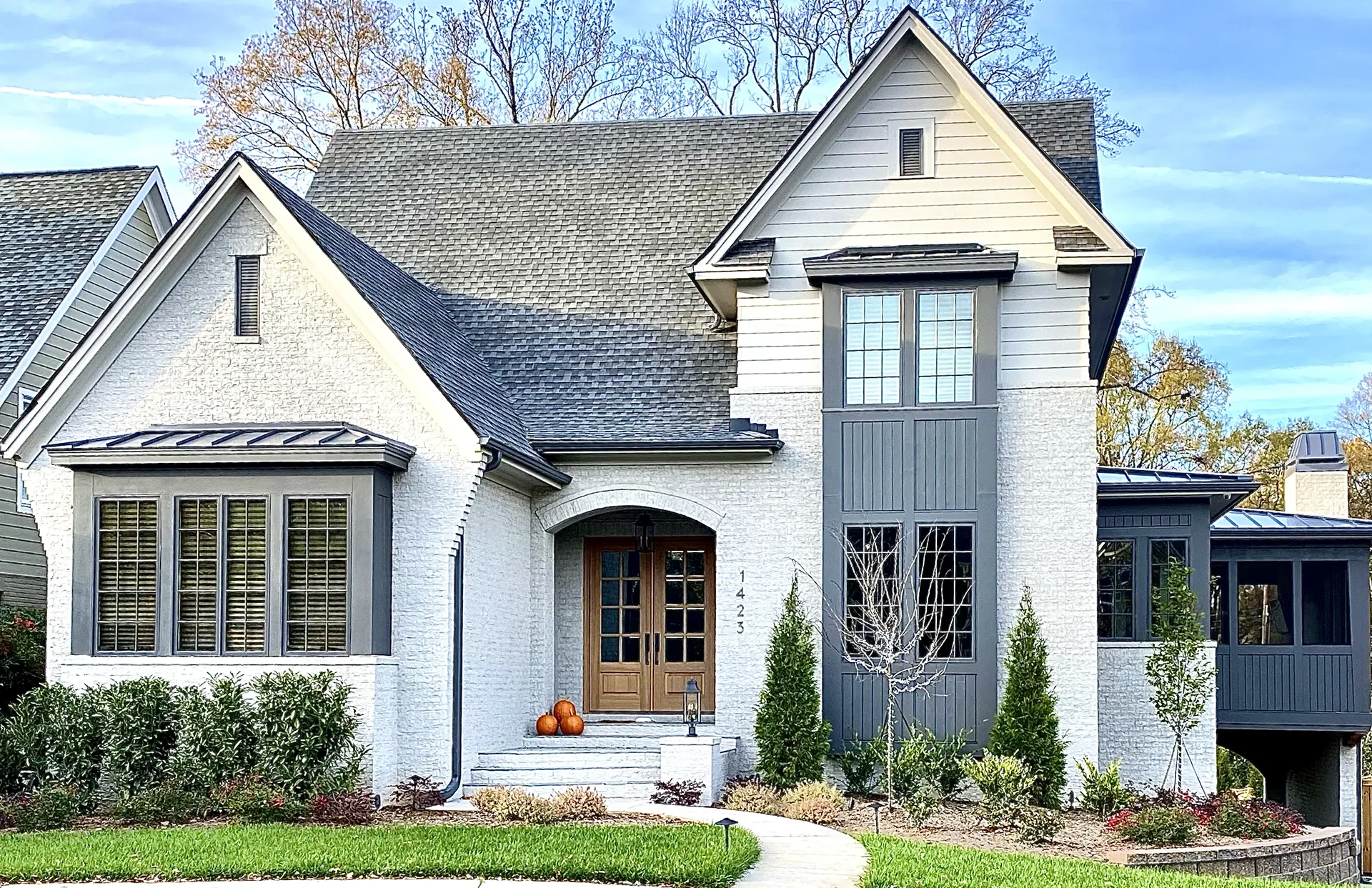 A stylish two-story home with white stucco and gray accents, featuring a wooden front door, pumpkins on the steps, and a well-manicured lawn.