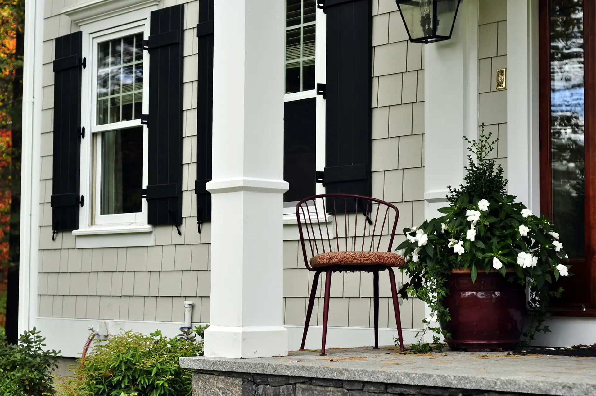 A red metal chair sits beside a potted white flower plant on a stone porch next to a house with black shutters.